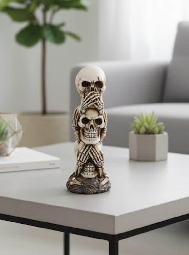 K&ouml;nnte beinhalten: Eine gestapelte Totenkopf-Figur in einer Pose von 'nichts sehen, nichts h&ouml;ren, nichts sagen'. Die cremefarbenen Sch&auml;del haben dunkle Augenh&ouml;hlen und halten sich die H&auml;nde vor Augen, Ohren und Mund. Die Figur steht auf einem grauen Tisch.