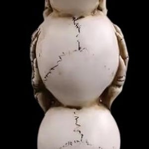K&ouml;nnte beinhalten: Eine gestapelte Skulptur aus drei Sch&auml;deln, jeder in einer anderen Position, mit H&auml;nden, die Augen, Ohren und Mund bedecken. Die Sch&auml;del sind cremefarben mit dunklen Linien und stehen auf einem strukturierten Sockel. Der Hintergrund ist schwarz.