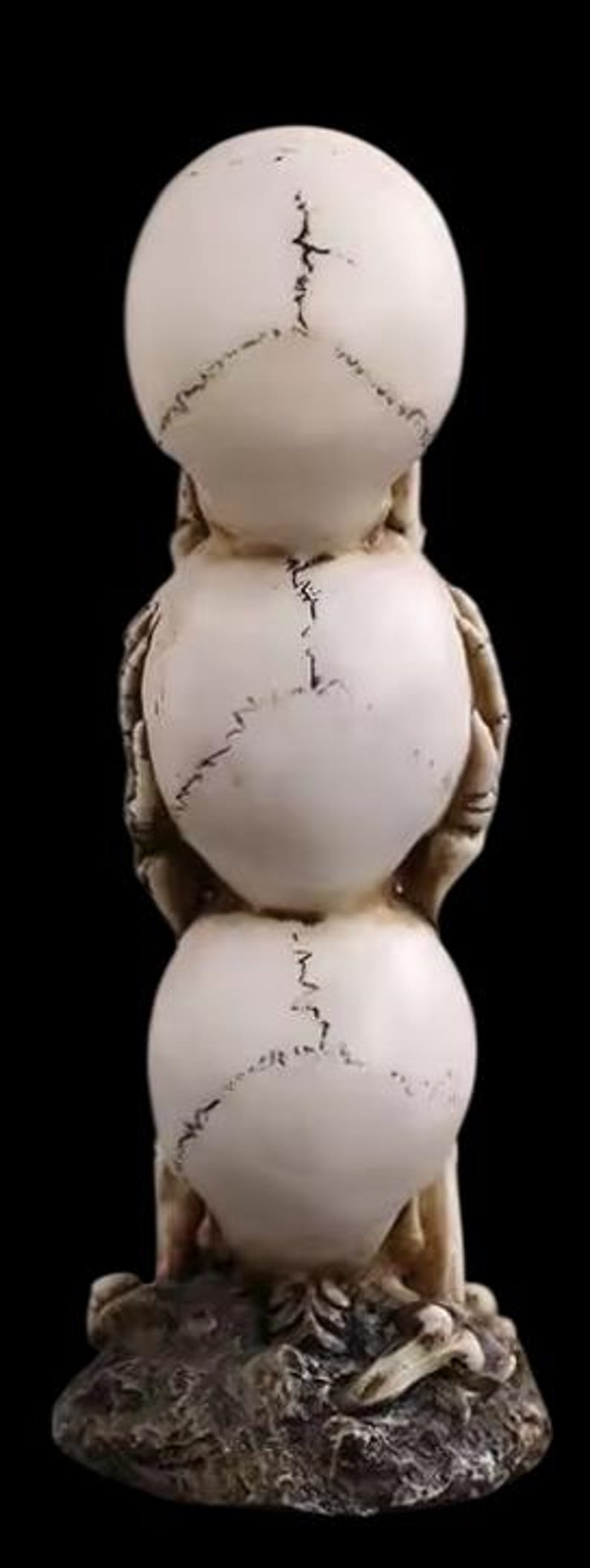 K&ouml;nnte beinhalten: Eine gestapelte Skulptur aus drei Sch&auml;deln, jeder in einer anderen Position, mit H&auml;nden, die Augen, Ohren und Mund bedecken. Die Sch&auml;del sind cremefarben mit dunklen Linien und stehen auf einem strukturierten Sockel. Der Hintergrund ist schwarz.