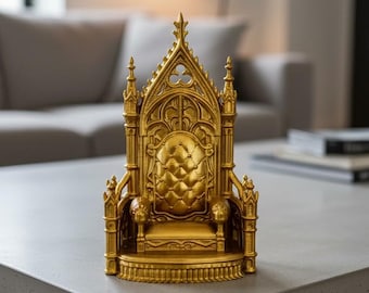 Puede incluir: Un pequeño trono dorado con intrincados detalles arquitectónicos. La silla presenta un respaldo alto con arcos apuntados, elementos decorativos y un asiento acolchado. El trono es un artículo decorativo, probablemente una pieza de colección o de exhibición.