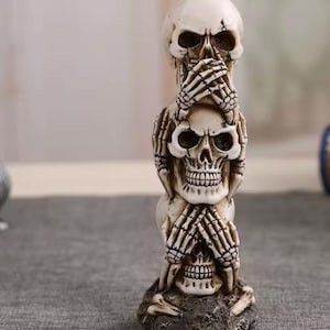 May include: A stacked sculpture of three skulls, each with a different hand gesture. The top skull has hands over its eyes, the middle skull has hands over its mouth, and the bottom skull has hands over its ears. The sculpture is off-white and sits on a dark, textured base.