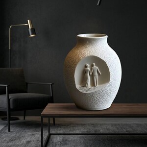 May include: A textured white ceramic vase with a circular cutout revealing a sculpted couple. The vase has a wide base and a narrow neck. The couple is depicted in a wedding scene. Home decor.