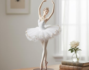 May include: A ballerina figurine in a graceful pose, wearing a white tutu with gold trim, a tiara, and pointe shoes. The figurine is mounted on a dark brown circular base. The ballerina's arms are raised, and her head is tilted.