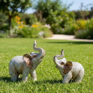 May include: Two decorative elephant figurines with raised trunks and detailed patterns. The elephants have brown ears and are set on a green lawn, with a garden in the background. The figurines are approximately 6 inches tall.