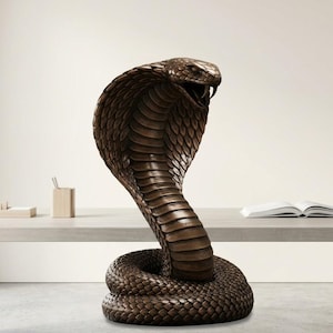 May include: A bronze cobra sculpture in a striking pose, with its hood expanded and mouth open. The sculpture has detailed scales and a coiled base. The cobra is a dark bronze colour, and the sculpture is set against a neutral background.