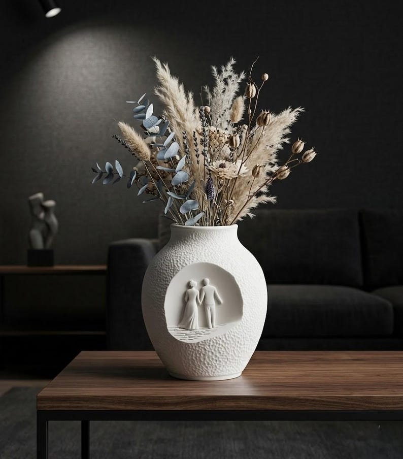 May include: A white ceramic vase with a textured surface and a carved relief of a couple. The vase holds a bouquet of dried flowers and pampas grass. The vase is on a dark wood table.