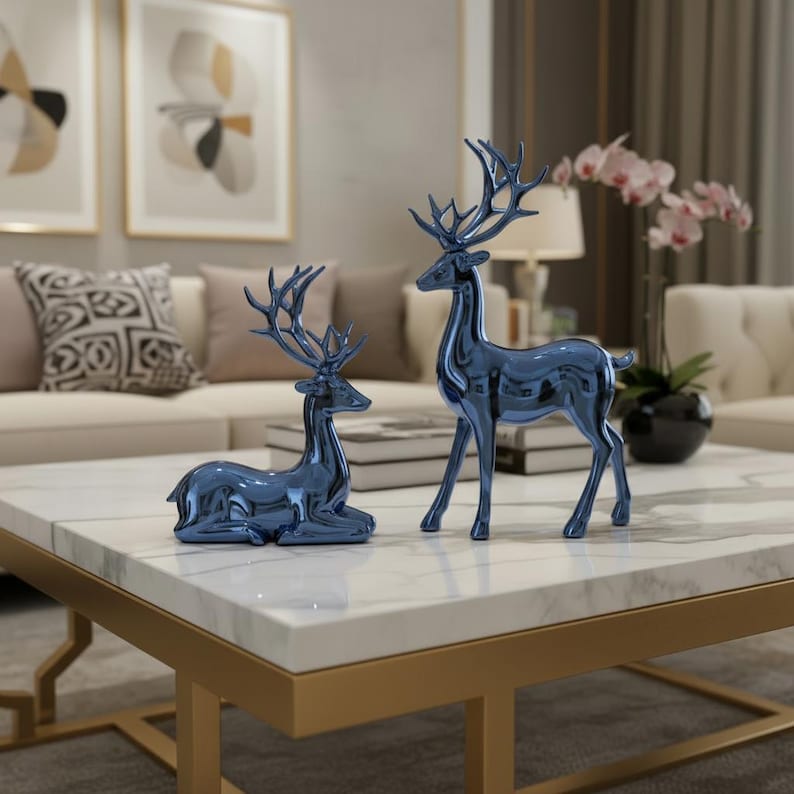 May include: Two blue decorative deer figurines. One deer is standing, the other is lying down. The figurines are placed on a white marble coffee table with gold-colored legs. The background includes a sofa, pillows, and artwork.
