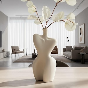 Op de afbeelding: Een crèmekleurige keramische vaas in de vorm van een vrouwelijk torso, met takken met witte bladeren. De vaas staat op een lichtgekleurde houten tafel, met een vage woonkamer op de achtergrond.