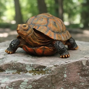 May include: A detailed, decorative turtle figurine with a textured, orange and brown shell, black legs, and a reddish-orange underside. The turtle is posed on a large, gray rock with moss, set against a blurred green background of trees.