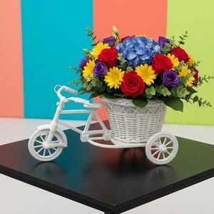 以下が含まれることがあります： 赤いバラ、黄色いデイジー、紫の花、青いアジサイが活き活きとアレンジされた、白い三輪車型のフラワーバスケット。バスケットは、カラフルな背景の黒いテーブルの上に置かれています。