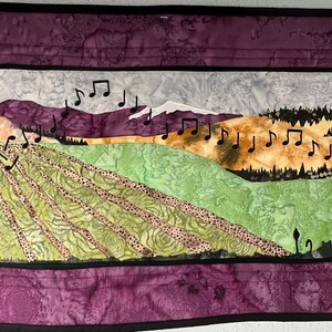 May include: A quilted wall hanging with a silhouette of a person blowing on a dandelion. The background features a landscape with mountains, fields, and a sky with musical notes.