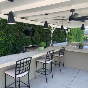 May include: Outdoor kitchen with a bar counter and four black metal bar stools with white cushions. The counter is made of concrete and has a built-in grill. The kitchen is surrounded by a green wall and a white pergola with a black ceiling fan.