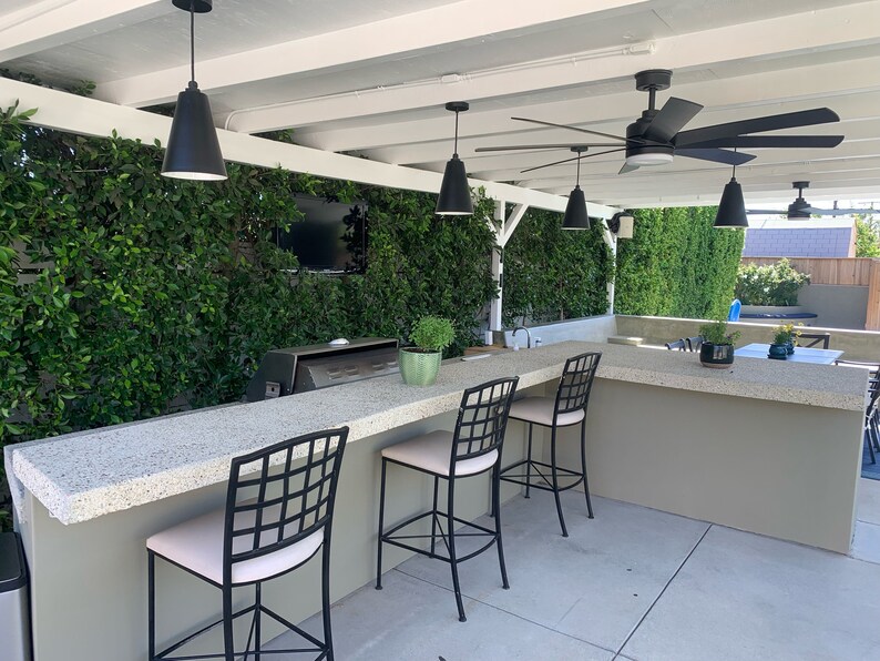 May include: Outdoor kitchen with a bar counter and four black metal bar stools with white cushions. The counter is made of concrete and has a built-in grill. The kitchen is surrounded by a green wall and a white pergola with a black ceiling fan.