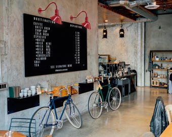 May include: Interior of a coffee shop with a black menu board, red lamps, and two bicycles. The menu lists coffee and tea options. A blue bicycle and a green bicycle are parked near the counter.