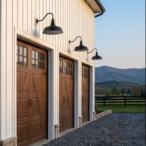 May include: Exterior view of a building with three dark brown wooden garage doors. Each door has a windowed top section. Black gooseneck lights are mounted above the doors. The building has white siding and a gravel driveway. Mountains are visible in the background.