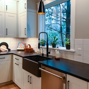 May include: A modern kitchen with white cabinets, black countertops, and a farmhouse sink. A black pendant light hangs over the sink. A stainless steel dishwasher is to the right of the sink. The window behind the sink looks out onto a wooded area.