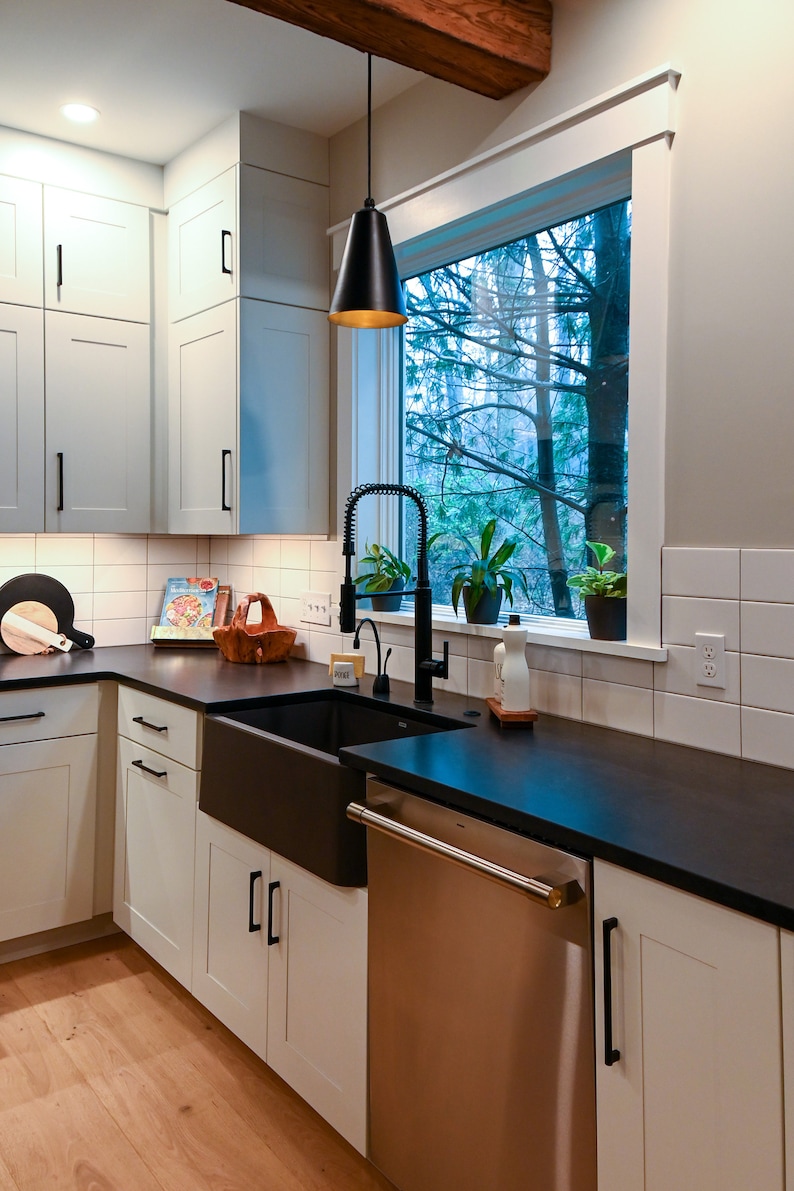 May include: A modern kitchen with white cabinets, black countertops, and a farmhouse sink. A black pendant light hangs over the sink. A stainless steel dishwasher is to the right of the sink. The window behind the sink looks out onto a wooded area.