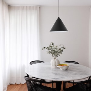 May include: A dining room with a round marble table and four black chairs. A black cone-shaped pendant light hangs above the table. A white vase with greenery and a bowl are on the table. Sheer white curtains frame the window.
