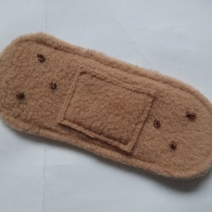 May include: A brown felt fabric bandage with a rectangular patch in the center. The bandage has several small brown stitches around the edges.