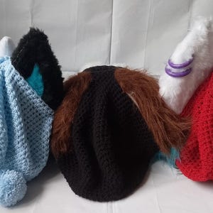 Könnte beinhalten: Drei gehäkelte Mützen in verschiedenen Farben: hellblau, schwarz und rot. Die Mützen sind mit einem einfachen Stichmuster gearbeitet und haben einen flauschigen Bommel oben.