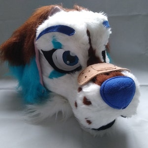 May include: A white and brown furry dog head with blue and pink accents. The dog has a blue nose and a brown patch on its eye. The head is made of soft, plush fabric and is suitable for use as a costume or prop.