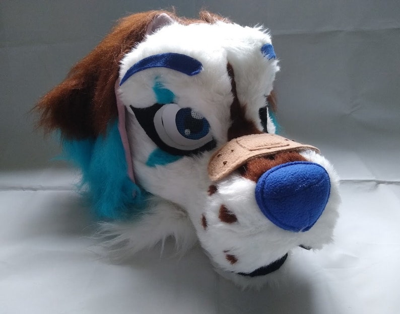 May include: A white and brown furry dog head with blue and pink accents. The dog has a blue nose and a brown patch on its eye. The head is made of soft, plush fabric and is suitable for use as a costume or prop.