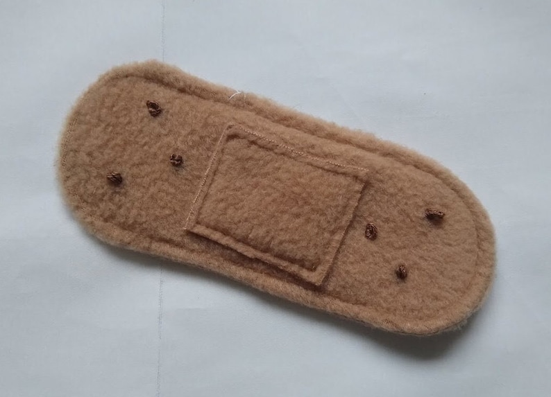 May include: A brown felt fabric bandage with a rectangular patch in the center. The bandage has several small brown stitches around the edges.