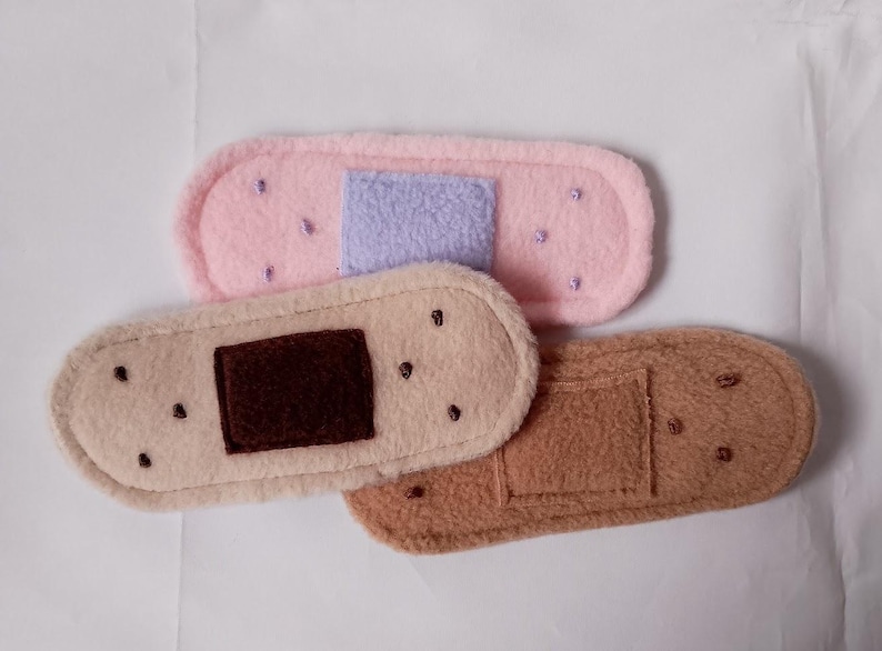 May include: Three plush fabric band-aid shaped toys. One is pink with a purple center, one is beige with a brown center, and one is brown with a brown center.