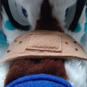 May include: Close-up of a brown plush toy with a brown felt patch sewn on. The patch has a square shape and is stitched with brown thread.