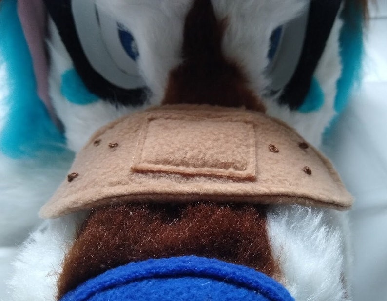 May include: Close-up of a brown plush toy with a brown felt patch sewn on. The patch has a square shape and is stitched with brown thread.