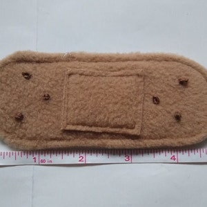 May include: A brown felt fabric bandaid with a square patch in the center. The bandaid is about 1.6 inches long and 1 inch wide.