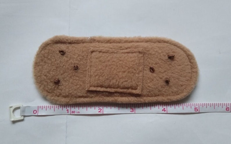 May include: A brown felt fabric bandaid with a square patch in the center. The bandaid is about 1.6 inches long and 1 inch wide.