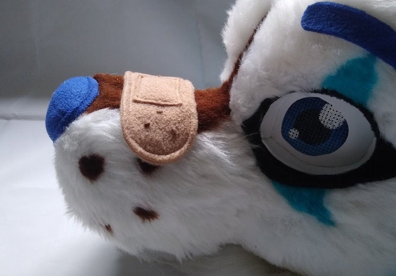 May include: White furry animal head with brown spots, blue and turquoise markings, and a brown patch with a tan bandage on the side. The head has a blue ear and a blue eye with a black mesh covering.