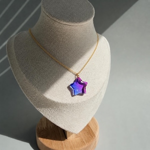 May include: A gold-tone chain necklace featuring a star-shaped pendant. The pendant is made of translucent material with a gradient color effect, transitioning from deep purple at the top to a vibrant blue at the bottom, catching the light.