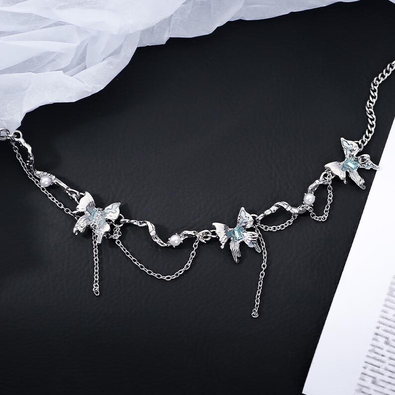 May include: A silver-toned necklace featuring three butterfly pendants with blue accents and dangling chains. The necklace has a delicate chain design with small pearl-like beads. The jewelry is displayed on a black surface with a white fabric in the background.