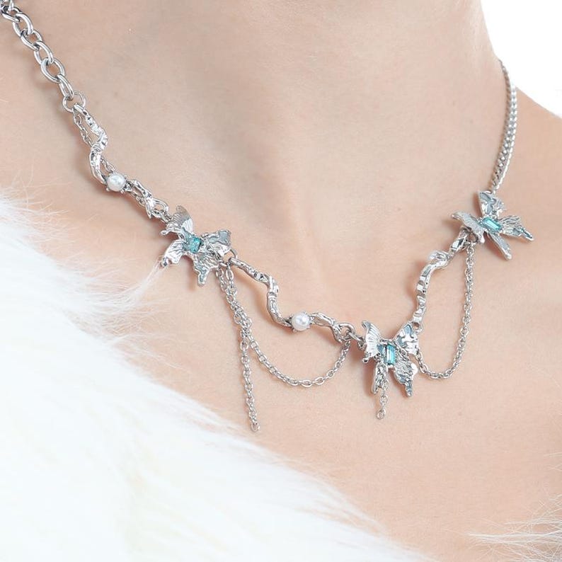 May include: Silver-toned necklace featuring a delicate design with butterfly and floral motifs. The necklace has small, light blue accents and dangling chain details. Small pearls are interspersed throughout the design, adding a touch of elegance.