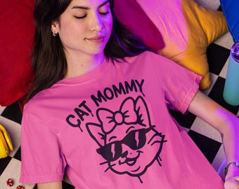 May include: Pink t-shirt with the text "CAT MOMMY" and a graphic of a cat wearing sunglasses. The cat graphic has a bow and a smiling face. The shirt is on a black and white checkered surface.