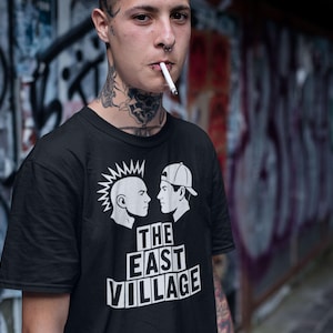 Puede incluir: Camiseta negra con un gráfico de dos perfiles laterales enfrentados, uno con cresta y otro con gorra de béisbol. El texto "THE EAST VILLAGE" está impreso debajo del gráfico en negrita.