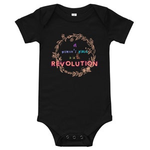 Puede incluir: Body de bebé negro con el texto "a womxn's place is in the REVOLUTION" en un diseño de corona floral. El texto es en tonos morados y rosas. El body tiene mangas cortas y corchetes en la parte inferior.