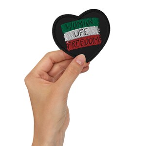 Könnte beinhalten: Ein schwarzer herzförmiger Aufnäher mit gesticktem Text und einem Flaggenmuster in Grün, Weiß und Rot. Die Worte "WOMAN LIFE FREEDOM" sind über die Flagge gestickt. Der Aufnäher wird in einer Hand gehalten.
