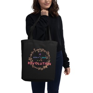 Puede incluir: Bolso tote negro con el texto "a womxn's place is in the revolution" en un diseño de corona floral. El bolso lo sostiene una persona que lleva una camisa negra de manga larga.