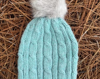 May include: A light mint green knit beanie featuring a vertical cable knit pattern throughout the body of the hat. The beanie is topped with a large, fluffy, light gray faux fur pom-pom that sits centered on the crown of the headwear.