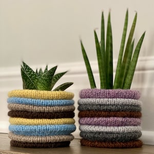 Op de afbeelding: Twee potplanten in gebreide stoffen potten. De plant links is een succulent met groene bladeren. De plant rechts is een slangplant met lange, groene bladeren. De potten zijn gemaakt van meerdere lagen gebreid stof in verschillende kleuren, waaronder geel, blauw, bruin, paars en grijs.