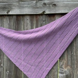 May include: A triangular knit shawl in a solid light purple color. The fabric features a repeating pattern of vertical stockinette stitch columns alternating with narrow, openwork eyelet lace stripes that run diagonally across the entire piece.