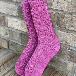 May include: A pair of pink knitted socks with a cable knit pattern. The socks are made with a soft, textured yarn.