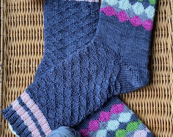 May include: A pair of hand-knitted socks in dark blue yarn. One sock features a textured diamond pattern with light pink stripes at the cuff. The other sock displays a colorful scalloped pattern in pink, green, white, and light blue.