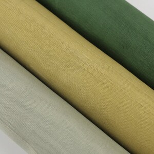 May include: Close-up of three rolls of fabric in shades of green, gold, and light grey. The fabric has a textured weave, and the rolls are angled diagonally. The colours are muted and natural.