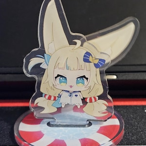 Pode incluir: Figura transparente em acrílico estilo anime com um personagem de desenho animado com orelhas grandes, olhos azuis e cabelo loiro. A figura está sobre uma base redonda com listras vermelhas e brancas, que se assemelha a um salva-vidas. O personagem usa uma roupa branca com detalhes em azul.