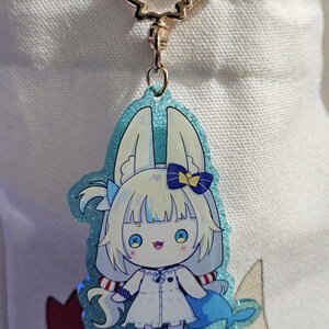 May include: A cartoon-style keychain in blue and white, depicting a character with white hair, blue eyes, and a blue bow. The keychain has a gold-coloured clasp and is attached to a white tote bag.