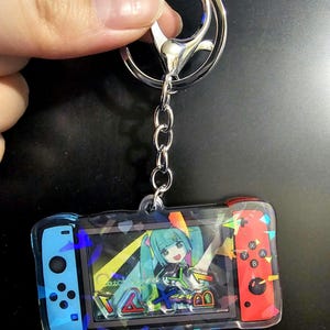 May include: A clear acrylic keychain with a video game controller design. The controller has blue and red sides and a character illustration in the centre. The keychain is attached to a silver metal clasp and chain.